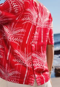 Rotes Kurzarmhemd mit weißem Palmenblattmuster, strukturierter Stoff und Knopfverschluss hinten.