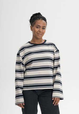 Jonge vrouw met gevlochten haar die lacht, draagt een beige en zwart gestreept shirt met lange mouwen en een zwarte broek, staand tegen een effen achtergrond.