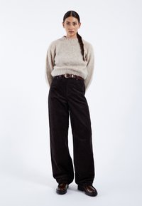 Jonge vrouw met lang gevlochten haar, die een beige trui draagt, brede zwarte broek met een bruine riem en donkerbruine schoenen, staand met handen achter de rug.
