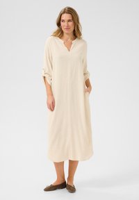 Beige, knälång klänning med en inskärning vid halsen, trekvartslånga ärmar och en lös passform. Inkluderar sidofickor för praktisk användning.