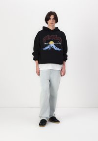 Svart oversized hettegenser med fjell- og solgrafikk, hvit lagdelt t-skjorte under, lyseblå jeans og svarte sneakers. Myk tekstur.