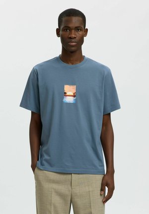 Jeune homme en t-shirt bleu avec un imprimé coucher de soleil et océan, portant un pantalon beige, debout devant un fond clair uni.