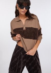 Maglione marrone lavorato a maglia con una striscia marrone pelosa e colletto henley; abbinato a pantaloni scuri in velluto con motivo a logo ripetuto.