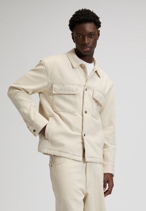 Junger Mann trägt eine elfenbeinfarbene, strukturierte Jacke und passende Hose, steht mit einer Hand in der Tasche vor einem schlichten grauen Hintergrund.