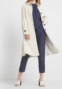 Kremowy płaszcz trencz z czarnymi guzikami, noszony na granatowym kombinezonie w kropki. Zawiera zaznaczoną talię, spodnie o długości kostek i białe sandały na obcasie.