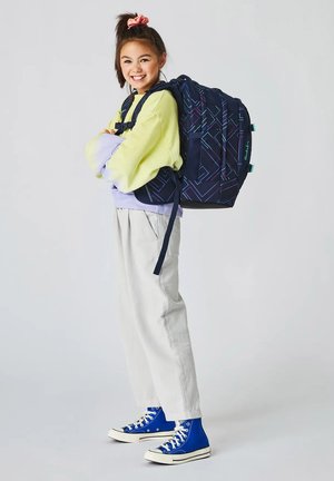 Lächendes Mädchen mit einem großen, gemusterten Rucksack, gelbem und lila Sweatshirt, heller Hose und blauen High-Top-Sneakern.