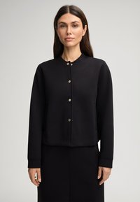Sweatshirt noir court avec un col rond, des manches longues et des fermetures à boutons métalliques. Tissu lisse avec des poches latérales.