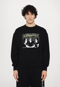 Black sweatshirt made of cotton with a relaxed fit. Features a graphic print in white and yellow depicting three characters.
