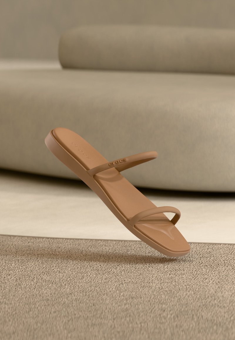 Καφέ σανδάλι τύπου slip-on με δύο λεπτές λουρίδες, τοποθετημένο πάνω από μια μπεζ υφασμάτινη επιφάνεια και μπροστά από ένα ελαφρώς μπεζ επενδυμένο φόντο.