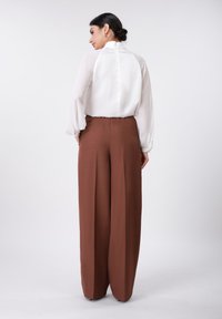 Blouse blanche à col montant et manches bouffantes, associée à un pantalon large marron sur mesure, vue de dos.