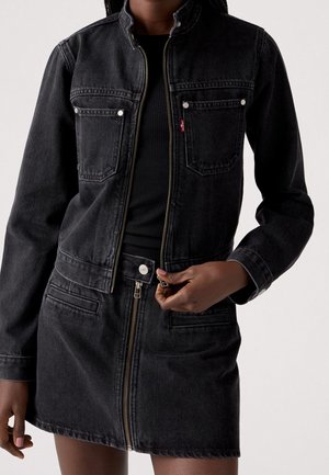 Femme portant une veste en jean noire et une jupe assortie avec des fermetures éclair à l’avant, ajustant la fermeture éclair de la jupe avec sa main droite.