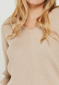 Beige gerippter Pullover mit V-Ausschnitt und vertikalen Nähten. Die Textur ist weich und hat einen subtilen Glanz sowie ein Wellenmuster.