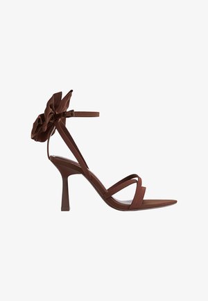 Brązowe sandały na obcasie z kwiatowym akcentem przy kostce. Posiadają cienkie paski, otwarty przód oraz zwężany kształt obcasa. Gładka faktura na całej powierzchni.