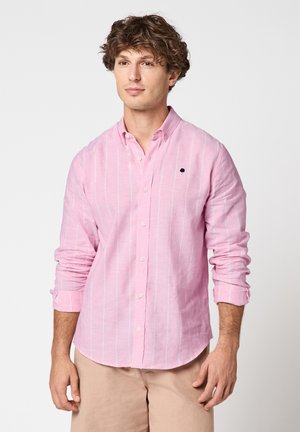 Junger Mann steht, trägt ein hellrosa gestreiftes Hemd mit Knöpfen und hochgekrempelten Ärmeln sowie beige Hose vor neutralem Hintergrund.