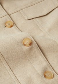 Manteau en laine beige avec trois boutons ronds marron clair, chacun à quatre trous, et des coutures le long du panneau avant. Texture douce.