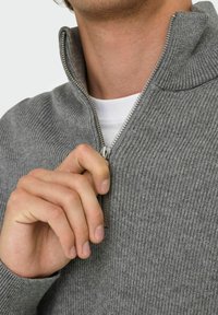 Grau zip-freier Pullover mit gerippter Textur, hohem Kragen und silbernem Metallreißverschluss, teilweise geöffnet, darunter ein weißes T-Shirt sichtbar.