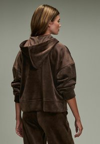 Braune Velours-Hoodie mit lockerer Passform, großer Kapuze und gerippten Bündchen. Strukturierter Stoff und subtiler Glanz verleihen ein weiches Aussehen.