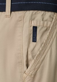 Beżowe szorty z tkaniny z teksturowanym ciemnoniebieskim pasem. Cecha to mała boczna kieszonka z ciemną metką oraz widoczne szwy.