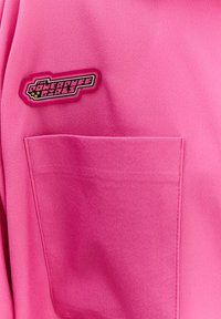 Ružová látková košeľa so širokou nášivkou s logom "Dievčatá z Powerpuff", prišitým vreckom a hladkou textúrou.