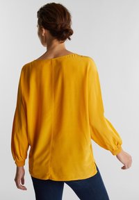 Blouse jaune avec une texture lisse et soyeuse, coupe ample et manches longues resserrées aux poignets. Le dos comporte une couture centrale pour la structure.