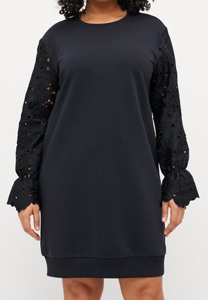 Vestido negro de manga larga con mangas de encaje calado con motivos florales y puños con volantes, llevado por una persona que está de pie frente a un fondo liso.