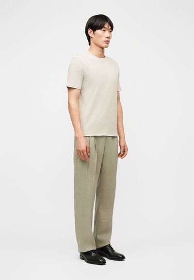 Άνδρας που στέκεται φορώντας ανοιχτό μπεζ κοντομάνικη μπλούζα, φαρδιά ελιά παντελόνια και μαύρα δερμάτινα παπούτσια φόρεμα, μπροστά σε απλό λευκό φόντο.