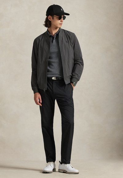 Mees musta nokamütsi, päikeseprillide, tumehall jaki, halli polo, mustade pükste, valgete jalanõude ja käekellaga, seisab neutraalse tausta ees.