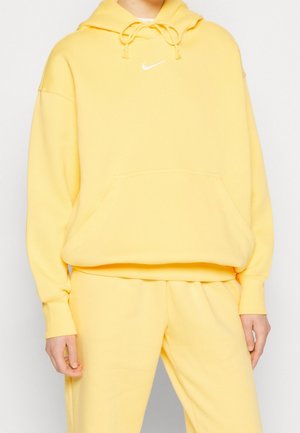 Osoba ubrana w pasujący żółty dres i bluzę Nike z kapturem, białe logo Nike widoczne na piersi, stoi na tle jednolitego jasnego tła.
