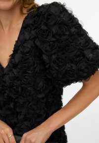 Melna kleita ar teksturētu auduma ziedu virsmu, dziļu V-veida izgriezumu un īsām piedurknēm, radot apjomīgu izskatu.