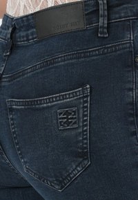 Sötétkék, slim fit farmernadrág, denim textúrával, és egy műbőr folttal, amin a "NOISY MAY" felirat található a hátsó zsebene. Finom varrási részletek.