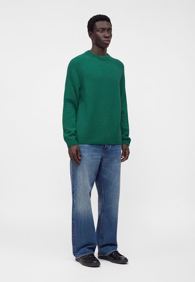 Homme debout portant un pull en maille vert, un jean bleu ample et des chaussures noires sur un fond blanc uni.