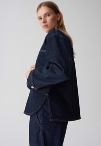 Giubbotto in denim, blu scuro, con cuciture a contrasto, maniche larghe e aperture laterali. Il tessuto ha una superficie liscia e un design strutturato.