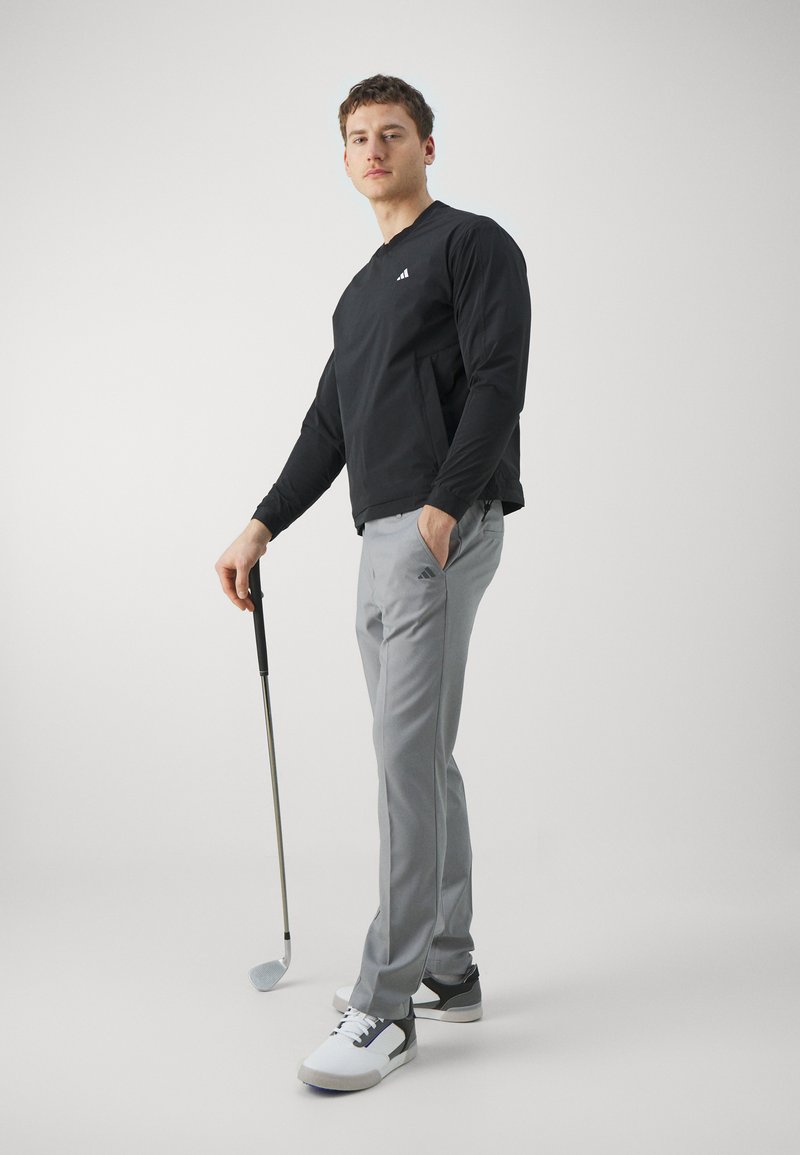 Schwarzes Langarmshirt mit weißem Logo, graue Golfhosen und schwarz-weiße Schuhe. Hält einen Golfschläger und steht vor einem schlichten Hintergrund.