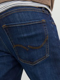 Mørkeblå denimjeans har en baklomme med et karakteristisk buet gult søm mønster. Stoffet har en jevn tekstur.