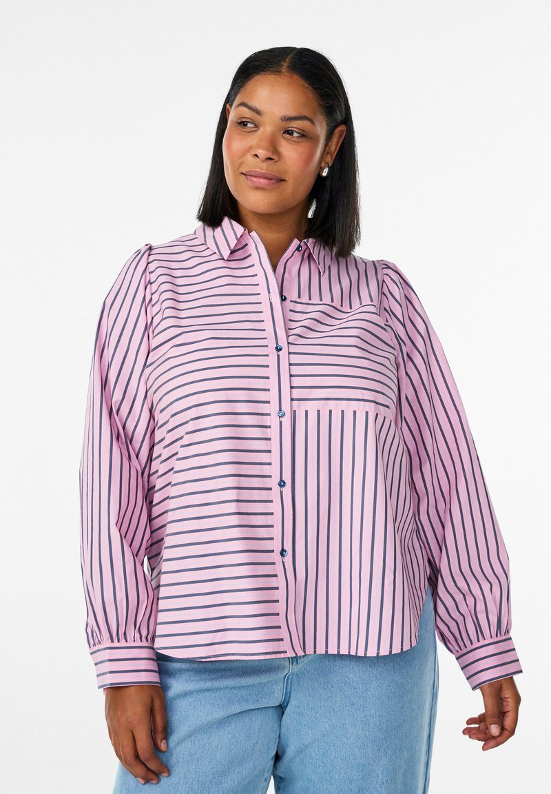 Nainen, jolla on yllään löysä vaaleanpunainen napitettava paita, jossa on pystysuoria ja vaakasuoria mustia raitoja, yhdistettynä vaaleansinisiin farkkuihin, seisomassa valkoisella taustalla.