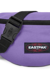 Fialová nylonová ledvinka s černým zipem. Má černý popruh a nášivku s logem Eastpak v černé a červené barvě. Texturovaný povrch.