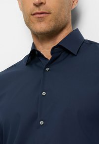Marineblaue Hemdbluse mit klassischem Kragen, glatter Textur und einer Knopfblende mit fünf Knöpfen in Silber. Dezentes Karomuster im Kragen.
