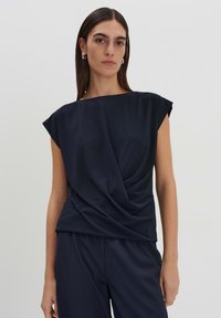 Top blu navy a maniche corte con design drappeggiato sul davanti. Tessuto liscio con vestibilità comoda e scollatura ampia; abbinato a pantaloni scuri.