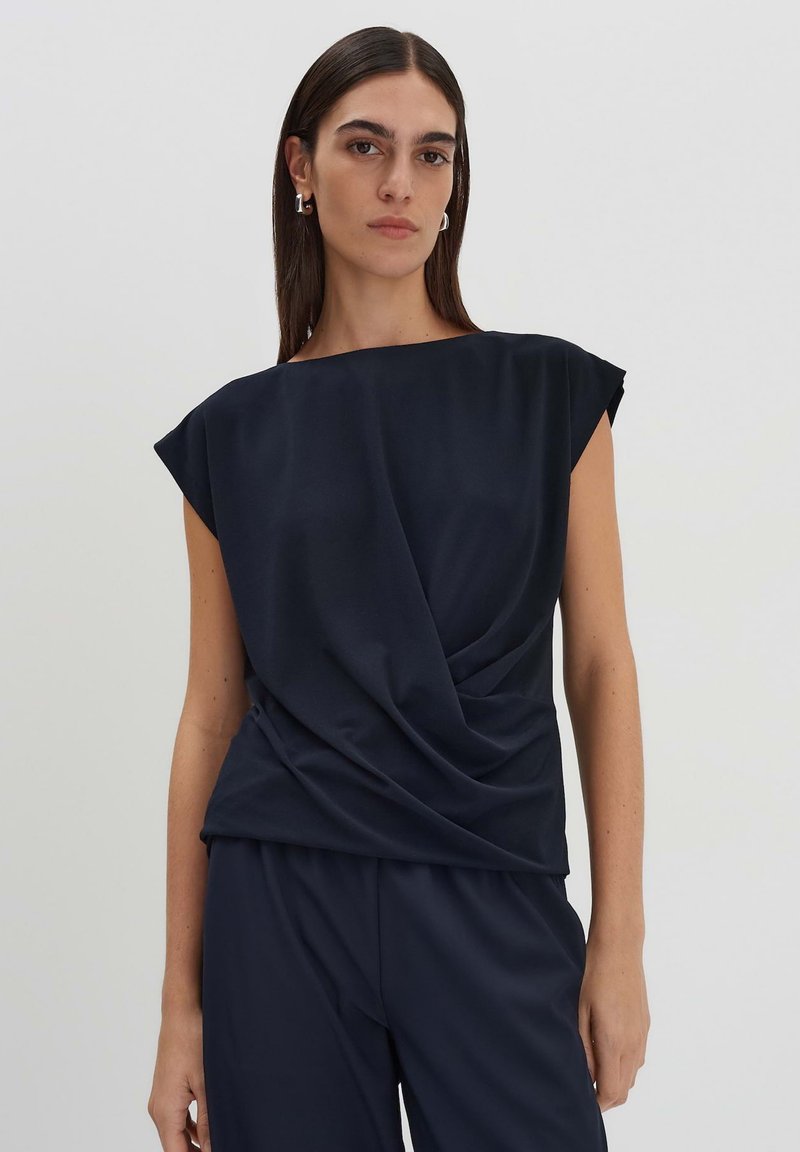 Top blu navy a maniche corte con design drappeggiato sul davanti. Tessuto liscio con vestibilità comoda e scollatura ampia; abbinato a pantaloni scuri.