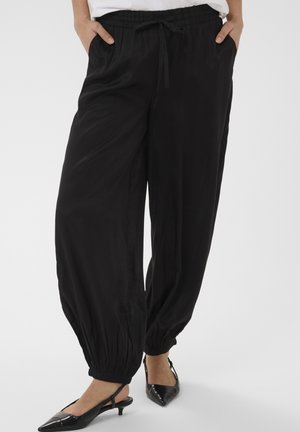 Femme portant un pantalon de jogging noir ample avec des poignets élastiques et des talons slingback noirs pointus, les mains dans les poches, sur fond blanc.
