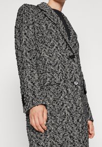 Schwarzer und weißer Blazer mit Fischgrätenmuster, zwei Knöpfen und Klappentaschen, der über einem schwarzen Oberteil getragen wird und einen Teil des Oberkörpers des Trägers zeigt.