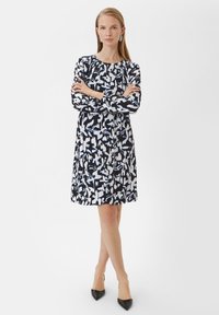 Langärmliges Kleid mit einem navyblauen und weißen abstrakten Blumenmuster, Rundhalsausschnitt und ausgestellt geschnittenem Saum. Kombiniert mit schwarzen spitzen High Heels.