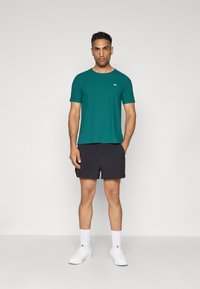 Türkise Crewneck-T-Shirt mit kurzen Ärmeln, kombiniert mit schwarzen Shorts. Das Model trägt weiße Sneakers und knöchellange Socken. Einfaches, lässiges Outfit.