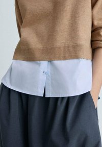 Brauner Strickpullover über einem weißen Hemd mit abgerundetem Saum, kombiniert mit einem dunkelgrauen, strukturierten Rock.