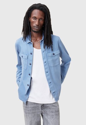 Light blue button-up shirt with two front pockets, long sleeves, and a soft texture, layered over a white tank top. Grey jeans visible.