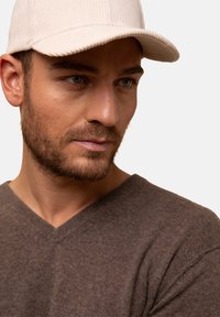 Béžová kordurová čepice s ribbovanou texturou, kterou nosí muž. Je oblečený v hnědém pleteném svetru s V výstřihem. Neutrální pozadí.