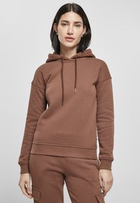Brun hettegenser laget av myk tekstil, med snorhette, ribbestrikkede mansjetter og en avslappet passform. Glatt tekstur med minimalt med detaljer.