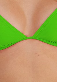 Heller grüner Bikini-Oberteil mit dreieckiger Form, glatter Textur und einer mittigen Naht, die die Cups verbindet. Einfaches Design ohne Verzierungen.
