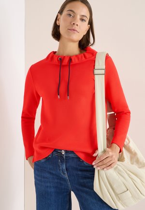 Rød pullover med bølget hætte og marineblå snøre, parret med blå denim jeans og en beige skuldertaske. Smoother tekstur, casual pasform.