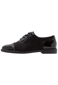 Scarpa nera da uomo con una combinazione di camoscio opaco e pelle verniciata lucida. Include lacci, punta arrotondata e piccoli dettagli decorativi.